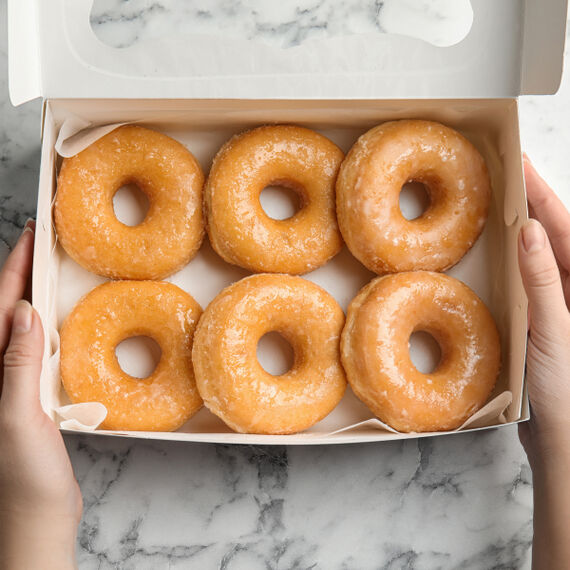 Zwei Hände halten sechs glasierte Donuts in einer Pappschachtel.