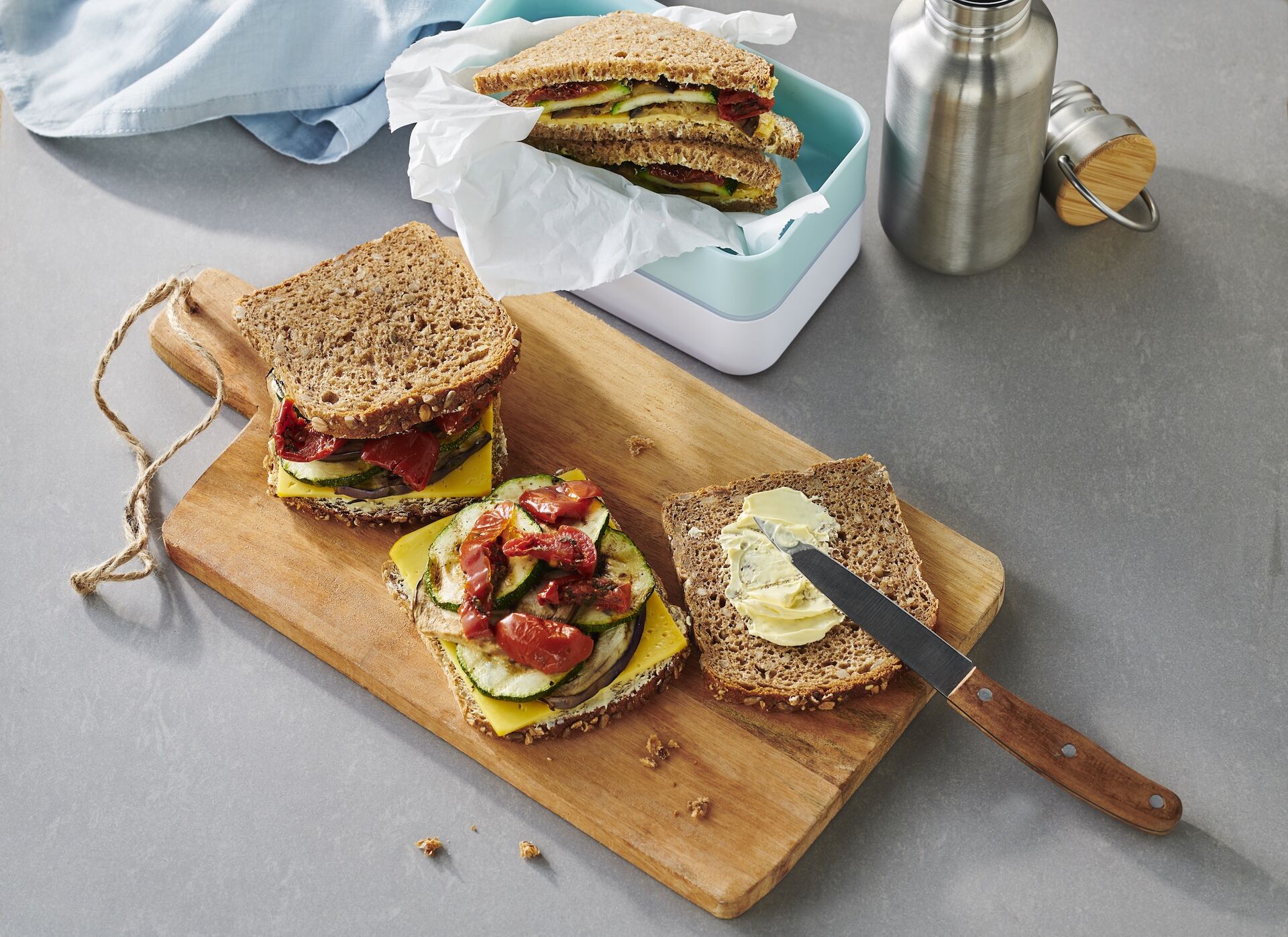 Auf einem Holzbrett und in einer Brotdose liegen Volle Pulle Stullen: mit Grillgemüse und Käse belegte Vollkornbrote.