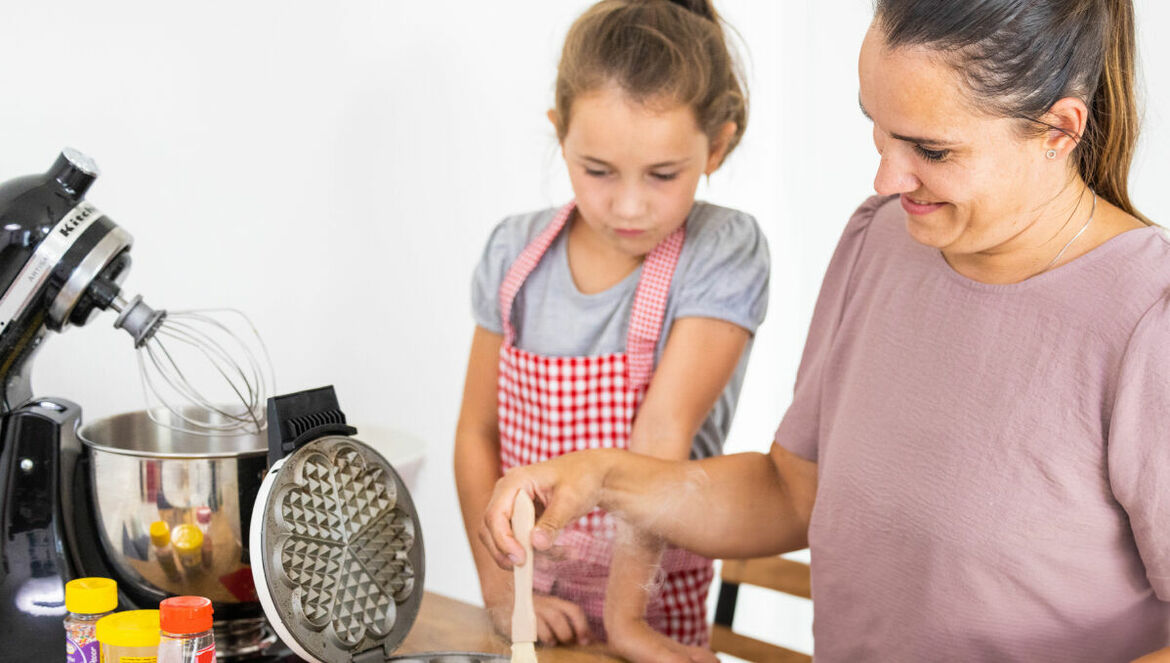 Eine Frau fettet ein Waffeleisen mit Margarine beim gemeinsamen Backen mit ihrer Tochter.