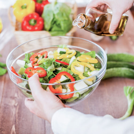 Ein Salat in einer Glasschüssel, die jemand in der Hand hält und Öl eingießt.