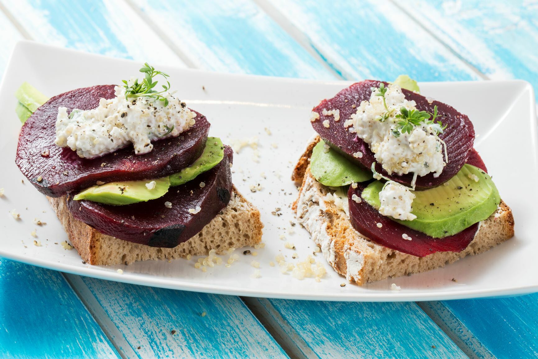 Auf einem weißen Teller liegen zwei halbe Superfood-Stullen belegt mit Roter Bete, Avocado und Quinoa.