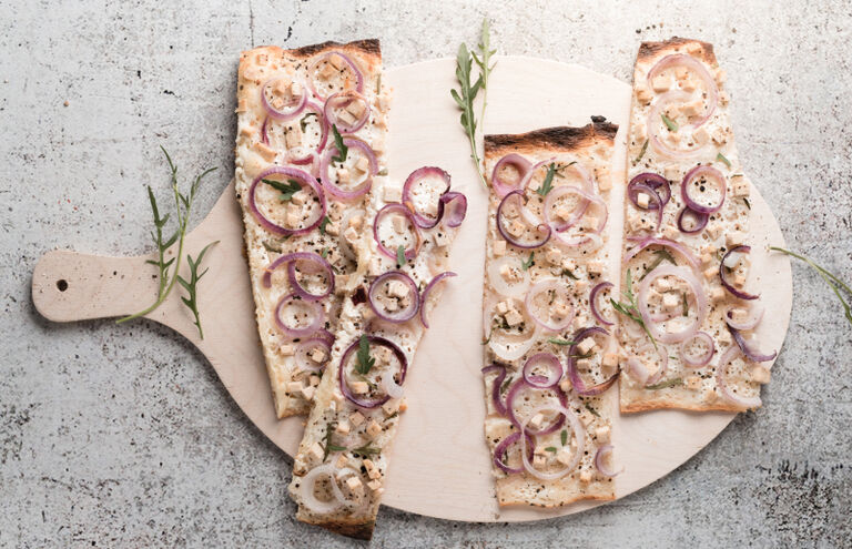 Vier Stücke Flammkuchen mit Zwiebelringen und Feta auf einem Holzbrett.