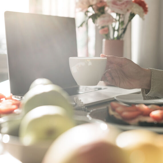 Eine Hand hält eine Kaffeetasse an einem Tisch, auf dem Obstteller, belegte Brote und ein offener Laptop stehen.