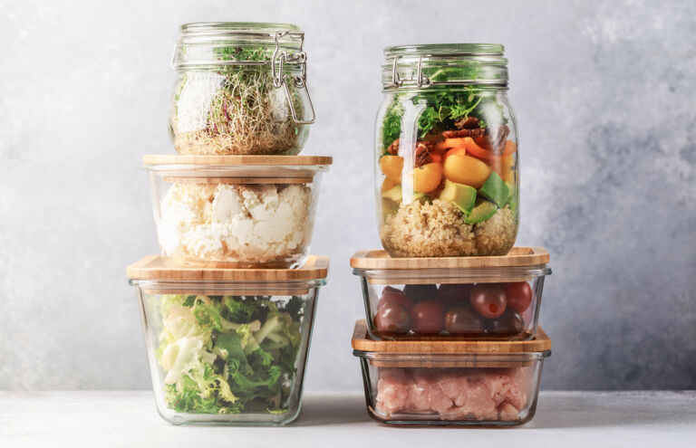 Gestapelte Vorratsdosen aus Glas, gefüllt mit portioniertem Salat, Obst und Fleisch.
