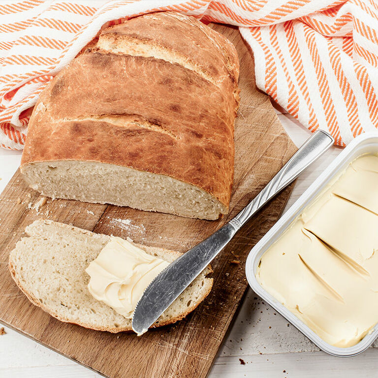 Eine abgeschnittene Scheibe Brot liegt vor einem Laib Brot auf einem Schneidebrett und wird mit einem Messer mit Margarine bestrichen