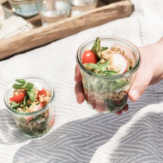 Hand mit Glas, das mit Salat gefüllt ist, daneben steht ein weiteres Salatglas auf einer Tischdecke.