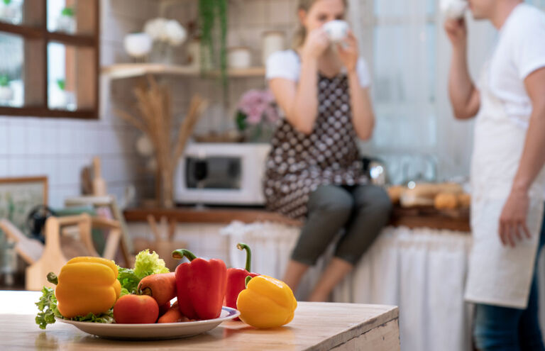 Ein Teller mit frischem Gemüse auf einem Küchentisch, im Hintergrund trinken ein Mann und eine Frau Kaffee.
