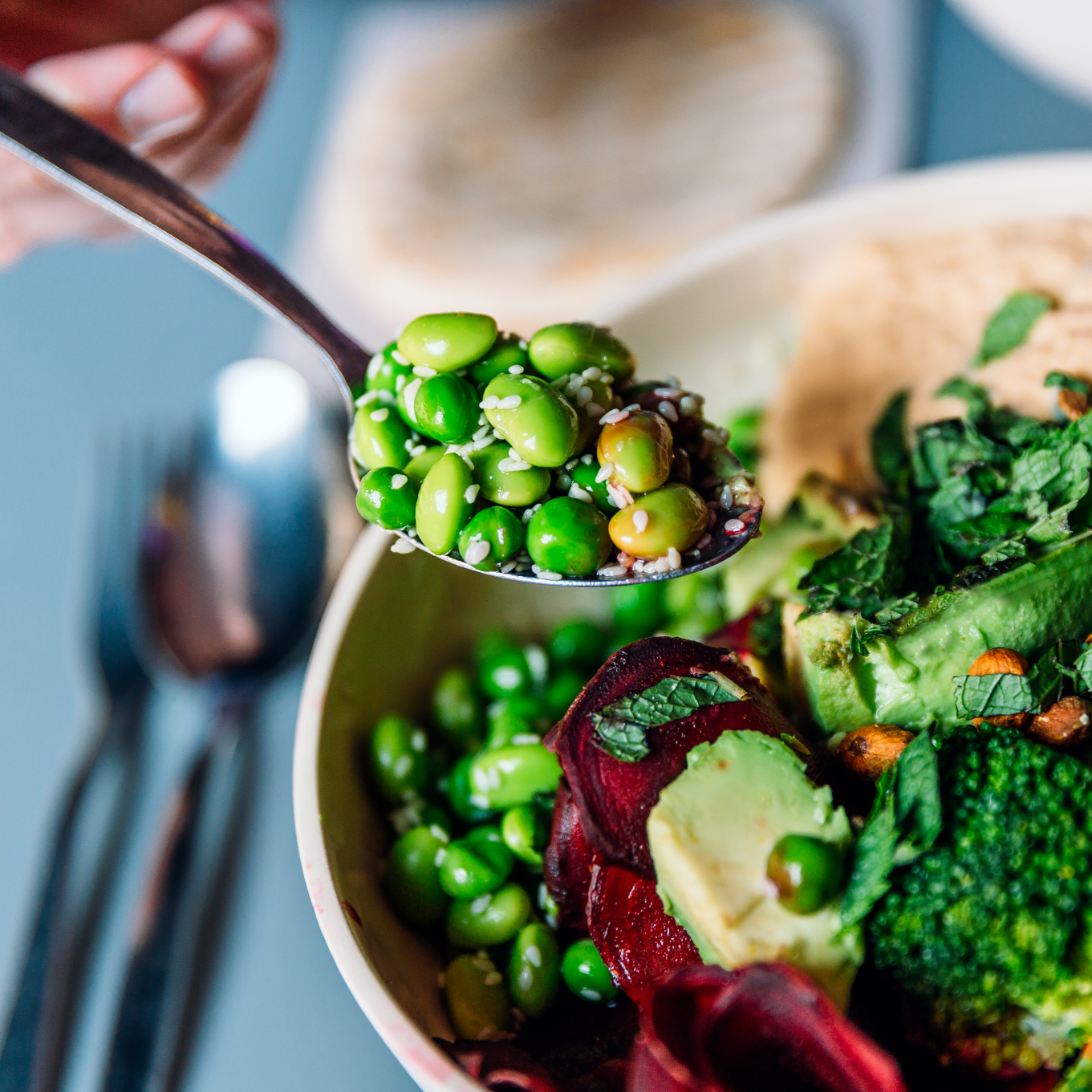 Eine Person isst eine vegane Bowl mit Edamame, Brokkoli, Avocado, Rote Bete, Hummus und Nüssen.